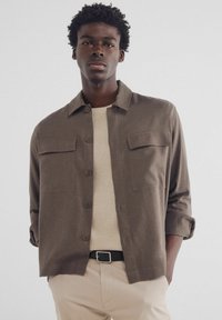 Lichtbruine blouse met een kraag, twee borstzakken en knoopsluiting, gedragen over een gestructureerde beige trui.