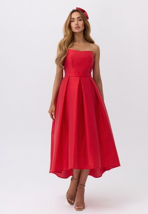 Kobieta o długich włosach, ubrana w czerwoną midi sukienkę bez ramiączek i pasującą opaskę, stojąca na białym tle.