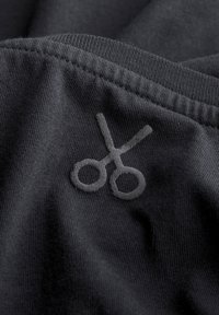 Zwarte stof met een gladde textuur en een tonale geborduurde schaarontwerp. De stof vertoont fijne ribbels en heeft een ontspannen val.