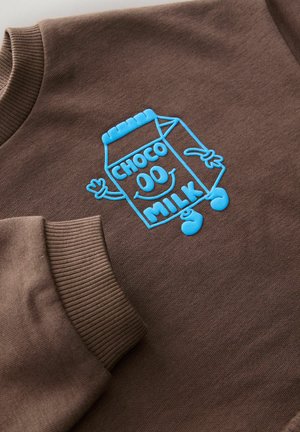 Sudadera marrón con un cartón de leche de dibujos animados bordado en azul, que lleva el texto "CHOCO OD MILK" y manos de dibujos animados, y puños acanalados texturizados.