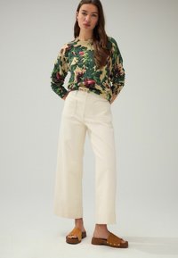 Een bloemen overhemd met lange mouwen in groen, roomkleur en bordeaux; roomkleurige wijde broek; bruine instapsandalen met houten hakken. Eenvoudige achtergrond.