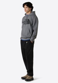 Grijze hoodie met zwart logo, voorzak, gedragen met zwarte broek en bruine wandelschoenen, met een robuuste textuur en veters.