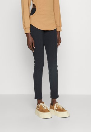 Ljusbrun långärmad topp med sidoklipp, i kombination med åtsittande svarta byxor och klumpiga bruna och vita sneakers. Enkel, modern design.