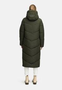 Lång pufferjacka i mörkgrön, med ett chevron-quiltat mönster, en huva och en etikett på baksidan. Bärs över vita byxor och boots.