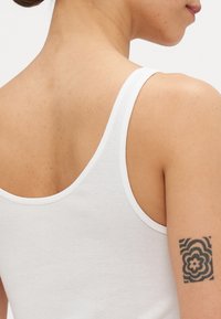 Hvit, stripe-mønstrete tanktop med tynne stropper. Mykt bomullsmateriale, tettsittende design. Inkluderer et synlig svart geometrisk tatovering på overarmen.