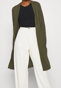 Oliivinvihreä ribbitetty neuletakki, jossa on avonainen etuosa, pitkät hihat ja sivutaskut, yhdistetty mustan paidan ja valkoisten leveälahkeisten housujen kanssa.