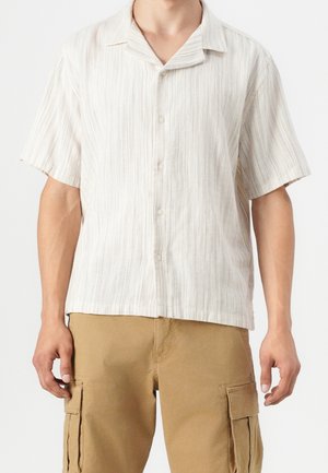 Mężczyzna ubrany w jasną, w paski koszulę z krótkim rękawem na guziki oraz beżowe spodnie cargo z bocznymi kieszeniami.