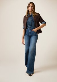 Tenue en denim comprenant une chemise ajustée avec col, une veste marron, et un jean large orné de clous en argent sur les côtés.