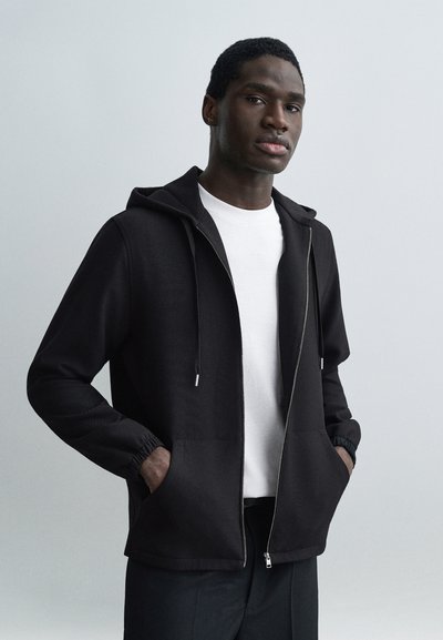 Jeune homme à la peau foncée portant un sweat à capuche noir zippé par-dessus un T-shirt blanc, les mains dans les poches, debout devant un fond uni gris clair.