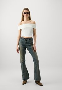 White off-shoulder crop top, blue flared denim jeans with faded texture, and brown pointed ankle boots, accessorized with black sunglasses.