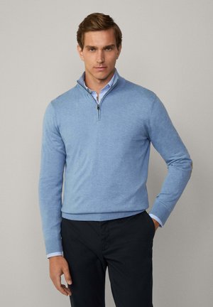 Suéter de cuello con cremallera en azul claro, hecho de una tela suave, con puños y dobladillo de punto acanalado, detalle de superposición en los codos, usado sobre una camisa con cuello.