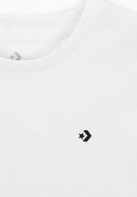 Camiseta de algodón blanca con cuello redondo, que presenta una pequeña estrella negra y un logotipo de chevron en el lado izquierdo. Textura suave.
