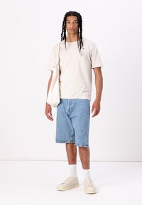 Tricou din bumbac bej cu logo, pantaloni scurți din denim albastru, șosete albe până la gleznă și adidași bej. Modelul ține o geantă de pânză albă.