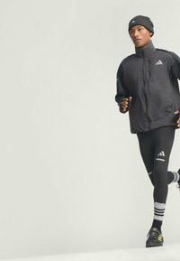 Black running jacket with high collar, black leggings with white stripes, and grey striped beanie. Running shoes with yellow accents.