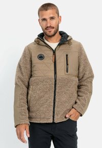 Tan og brun jakke, der kombinerer quiltet stof og tekstureret fleece. Har en lynlåslomme på brystet, justerbar hætte og et logo patch.