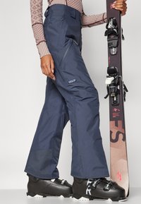 Ναυτικό μπλε παντελόνια σκι με χαλαρή εφαρμογή, που διαθέτουν πλαϊνούς φερμουάρ και λογότυπο. Μαύρες μπότες σκι με ασημί λεπτομέρειες και ένα χιόνι.