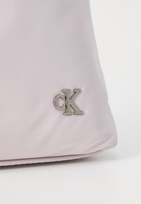 Borsa in tessuto lilla chiaro con una texture liscia e un piccolo accento del logo "CK" in argento sul lato. Design semplice e minimalista.