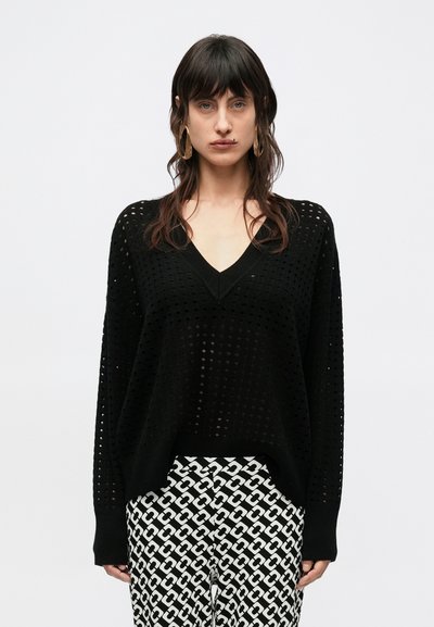 Pull noir en maille avec un col en V et un motif en treillis ouvert. Associé à un pantalon noir et blanc à motif, présentant un motif de maillons de chaîne.