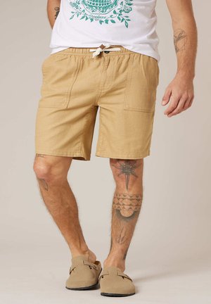 Homme portant un short beige avec cordon de serrage, des chaussures beige à enfiler, un t-shirt blanc avec un motif vert, et des tatouages visibles sur les jambes et les bras, debout sur un fond clair.