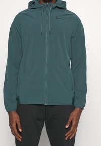 Hombre vistiendo una sudadera con cremallera color verde azulado, con cordones y el logo negro de Nike en el pecho, combinada con pantalones negros, de pie contra un fondo blanco.