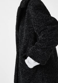 Manteau noir en peluche avec une finition texturée, doté d'un col structuré et de grandes poches latérales. Manches blanches retroussées visibles à l'ouverture.