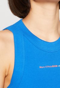 Blå ärmlös topp i ribbat tyg, med rundad halsringning och vit text med varumärket: "Marc O'Polo DENIM x."