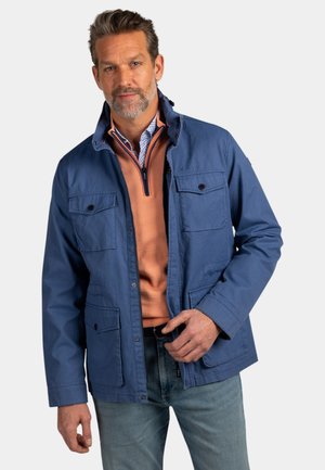 Man draagt een blauwe jas over een perzikkleurige trui met een kwartritssluiting en een overhemd met patroon, poseert terwijl hij met zijn hand de jas ter hoogte van de taille aanpast.