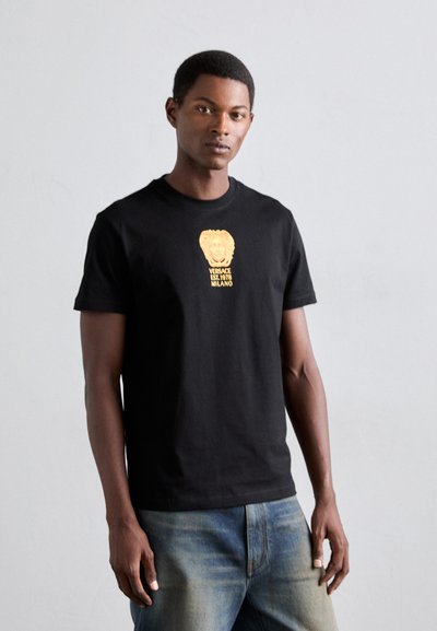Jeune homme portant un t-shirt noir Versace avec un logo et un texte dorés, associé à un jean bleu clair, se tenant devant un fond uni clair.