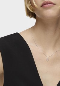 Collar de plata con un colgante simple y redondeado. La cadena es delicada y entrelazada, contra una blusa negra con escote en V.