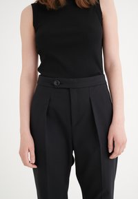 Top sans manches noir côtelé associé à un pantalon noir taille haute présentant un bouton unique et des plis marqués, créant un look habillé.