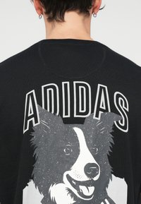 Černé bavlněné tričko s velkým grafickým potiskem bílého a šedého psa na zádech, s textem "ADIDAS" nahoře v tučných, kontrastních písmenech.