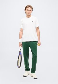 Νέος άνδρας με λευκό μπλουζάκι και πράσινο παντελόνι κρατώντας ρακέτα και μπάλα του τένις, στέκεται μπροστά σε απλό φωτεινό φόντο.