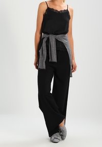 Camiseta sin mangas negra con encaje, suéter de punto gris anudado en la cintura, pantalones de pierna ancha negros y zapatos de cuadros tipo slip-on. Textura suave.