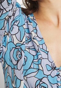 Blaues Stoffmuster mit floralem Design und leichter Textur, mit schwarzen Umrissen und subtilen goldenen Akzenten, im V-Ausschnitt-Design.