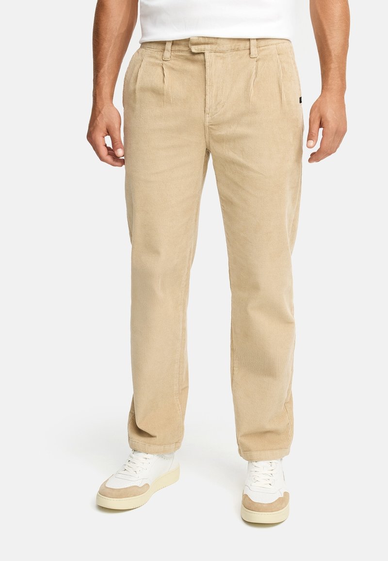 Homme portant un pantalon en velours côtelé beige et des baskets blanches, debout devant un fond blanc uni.