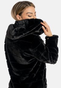 Giacca in ecopelliccia nera con cappuccio, texture trapuntata e polsini arrotolati. Il materiale è morbido e peloso, con dettagli di cucitura visibili.