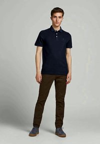 Νεαρός άνδρας που στέκεται μπροστά σε απλό φόντο φορώντας ναυτικές μπλούζες πόλο, καφέ παντελόνια και γκρι αθλητικά παπούτσια.