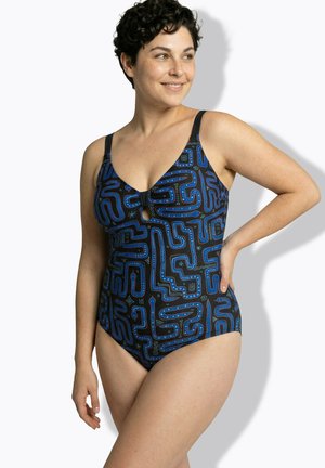 Kobieta z krótkimi kręconymi włosami, ubrana w czarny jednoczęściowy strój kąpielowy z niebieskimi abstrakcyjnymi wzorami przypominającymi labirynt, uśmiechająca się i patrząca na bok.
