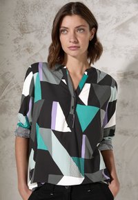 Blouse à imprimé géométrique multicolore avec des formes noires, teal, violettes et blanches, dotée d'un col en V et de détails de boutons. Texture de tissu douce.