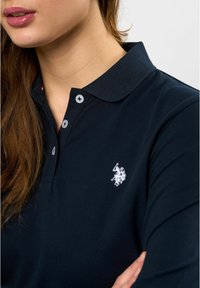 Marineblå langermet poloskjorte med krage, med to grå knapper og en hvit brodert logo på venstre bryst. Glatt tekstur.