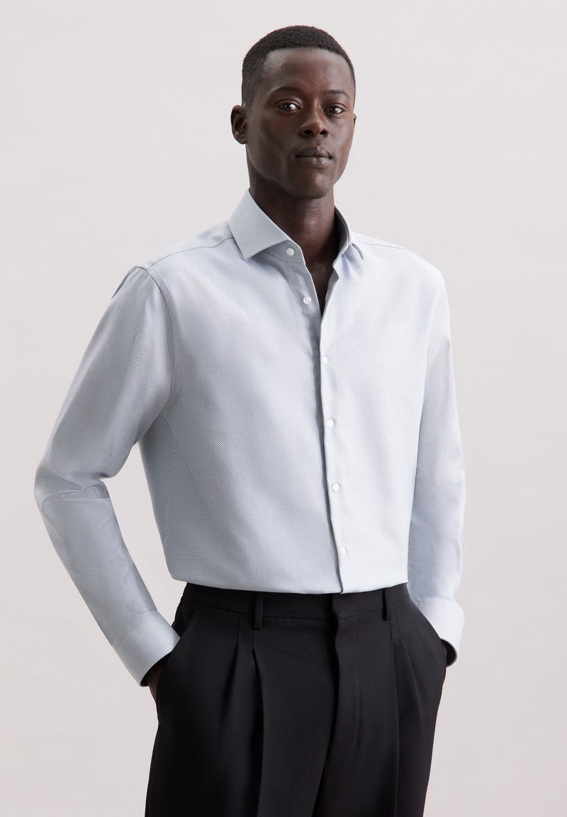 Chemise bleu clair à boutons avec un motif à carreaux subtil, à manches longues, coupe ajustée et col pointu, associée à un pantalon plissé foncé.
