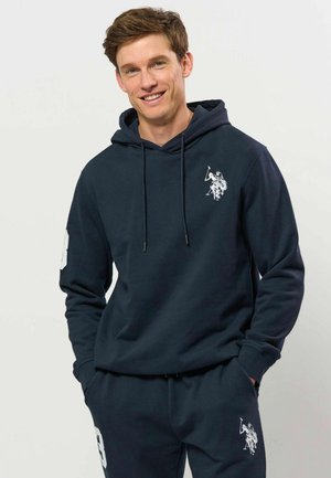 Jeune homme souriant portant un sweat à capuche bleu marine et un pantalon de jogging assorti avec des logos blancs de joueur de polo, mains dans les poches, sur un fond clair uni.