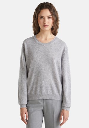Jeune femme aux cheveux bruns ondulés portant un pull léger gris clair à col rond et un pantalon gris clair, se tenant contre un fond blanc.