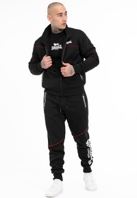 Zwarte zip-up jas en sweatpants met rode accenten. Tekstlogo op de borst en broek. Effen zwart shirt eronder. Witte sneakers.