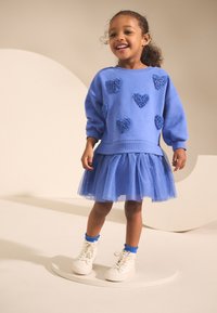 Kék pulóver szívapplikációkkal, réteges tüll szoknya és fehér tornacipő, kék szegélyű zoknival. Puha anyag, laza kialakítás.