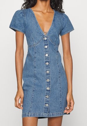 Kvinde iført en kortærmet blå denimkjole med dyb V-hals og knapper foran fra top til bund.