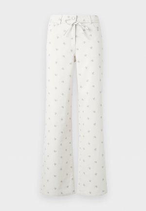 Mladá žena s krátkymi kučeravými blond vlasmi, oblečená v svetložltom tričku s dlhým rukávom, bielych širokých nohaviciach s malými vzormi a čiernych balerínach.