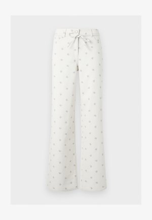 Νέα γυναίκα με κοντά σγουρά ξανθά μαλλιά, που φορά μπλούζα μακρυμάνικη σε απαλό κίτρινο χρώμα, λευκά παντελόνια φαρδιά με μικρά σχέδια και μαύρα φλατ παπούτσια.