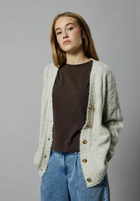Cardigan lavorato a maglia grigio chiaro con un motivo testurizzato, bottoni in legno e maniche larghe, abbinato a una camicia marrone scuro e jeans blu.