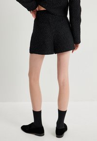 Pantalones cortos negros hechos de una tela texturizada con chispas metálicas, con una cintura alta y un dobladillo limpio. Combinados con calcetines y zapatos negros.
